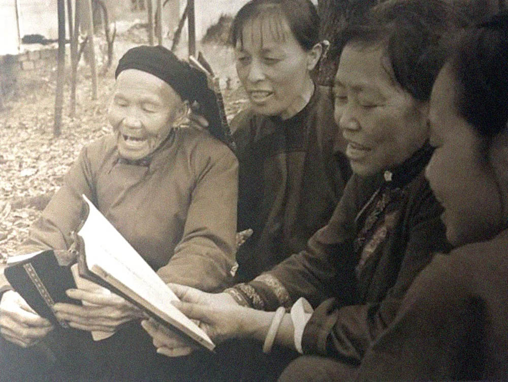 Photograph of four women singing and reading songs around a book written in Nüshu