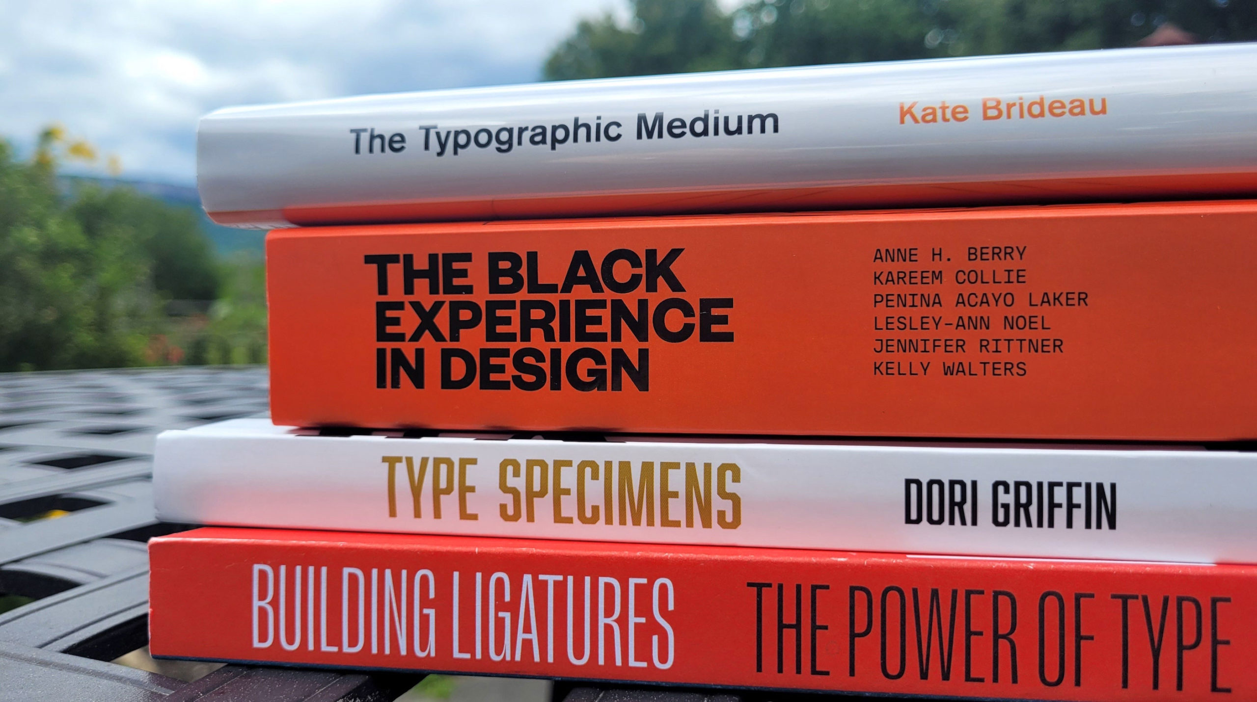 four books stacked on a table outside with some yellow flowers and greenery in the background. The books are from top to bottom: The Typographic Medium, The Black Experience in Design, Type Specimens, and Building Ligatures The Power of Type