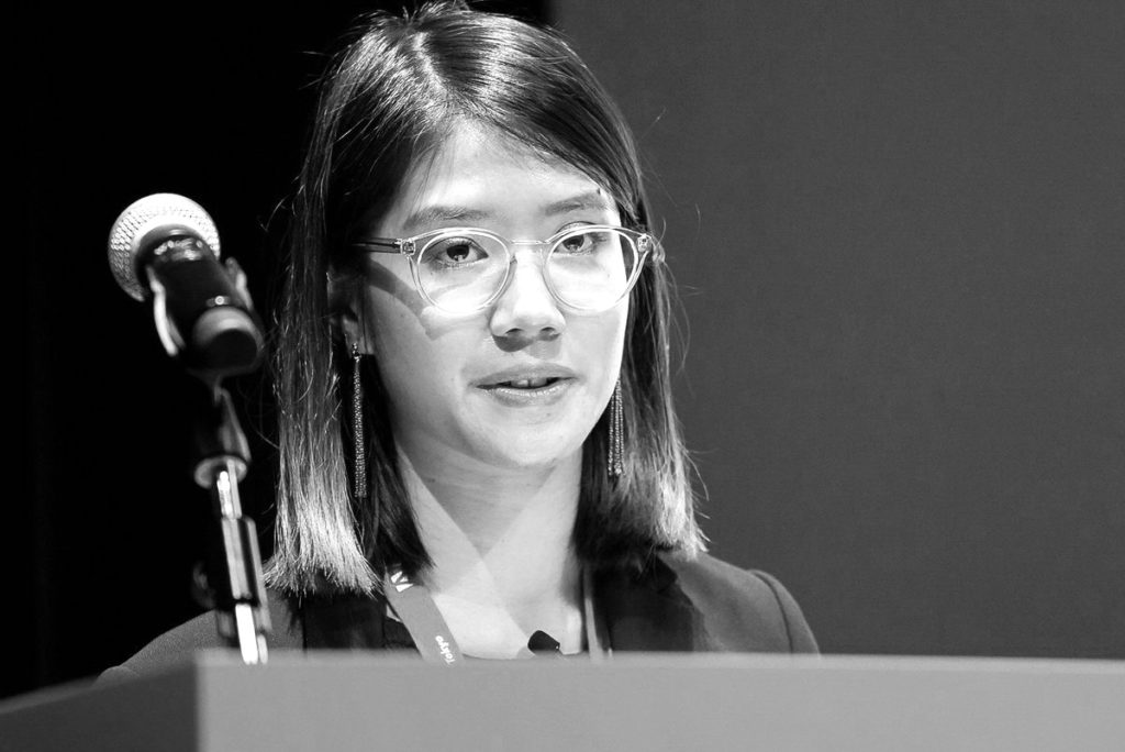 black and white headshot of a woman speaking at a podium with a microphone