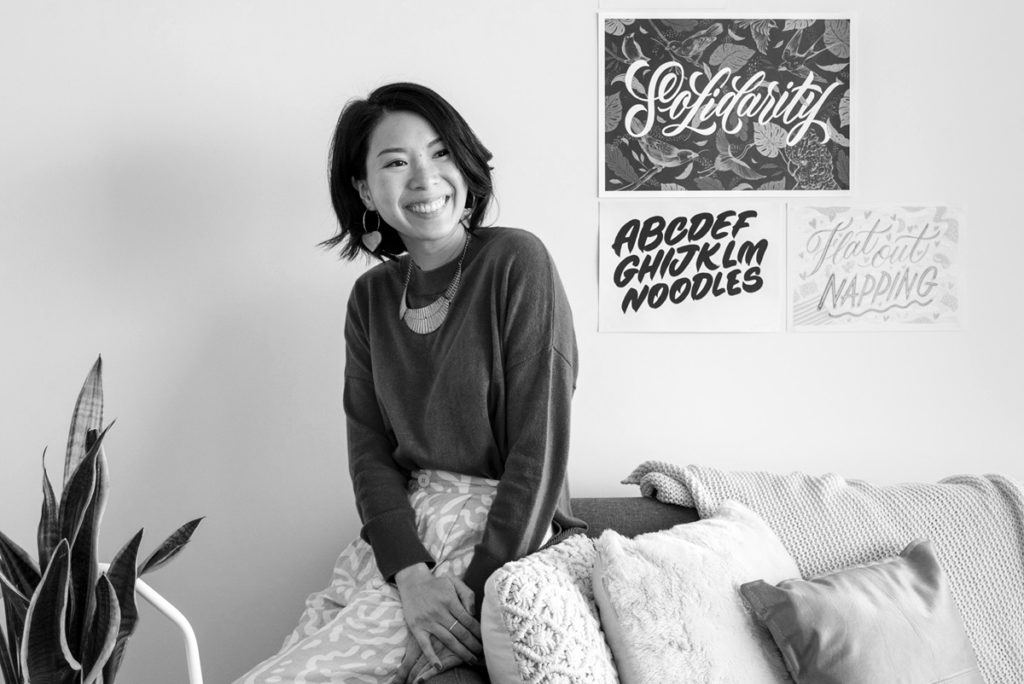 black and white photo of a woman smiling while leaning against a couch with lettering artwork on the wall