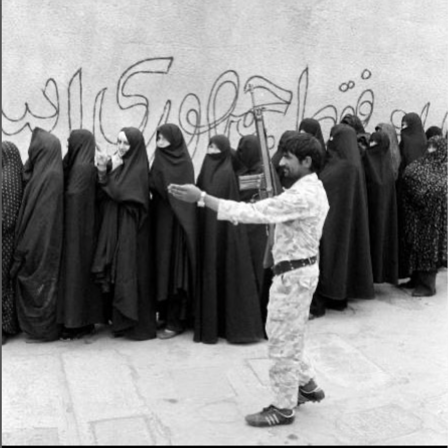 A male soldier stands in front of a line of women dressed in black covering