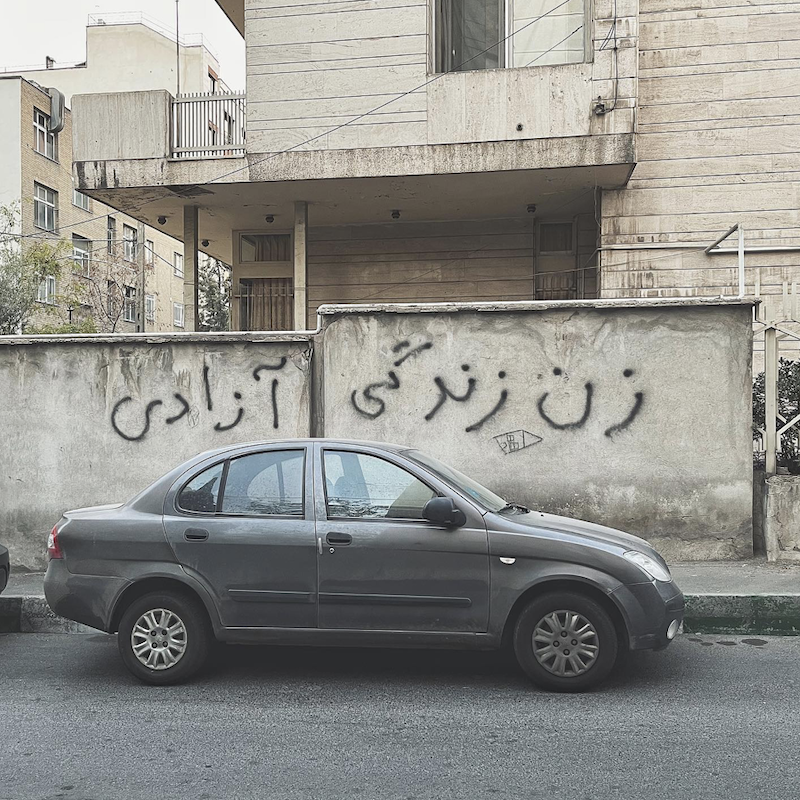 handwritten lettering on a wall next to a sidewalk with a small grey car parked on a street