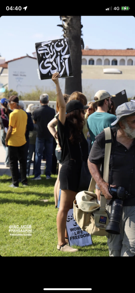 screenshot of an instagram story of a woman dressed in a black tshirt and shorts holding up a black sign with white lettering in a crowd of people