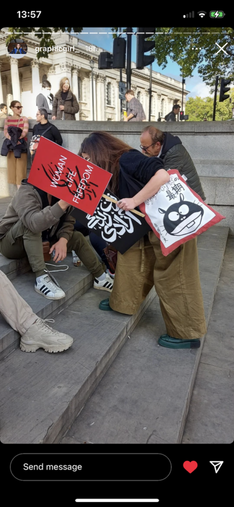 screenshot of an Instagram story of a woman leaning over steps at a rally holding a red sign with black and white lettering, a black sign with white lettering and a tote bag