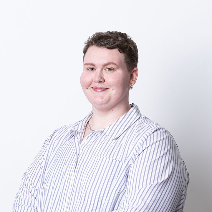 Image of Brooke Hull facing the camera at a 3/4 profile and smiling. They are a fat, white person with short, brown hair wearing a button up blue and white striped shirt.