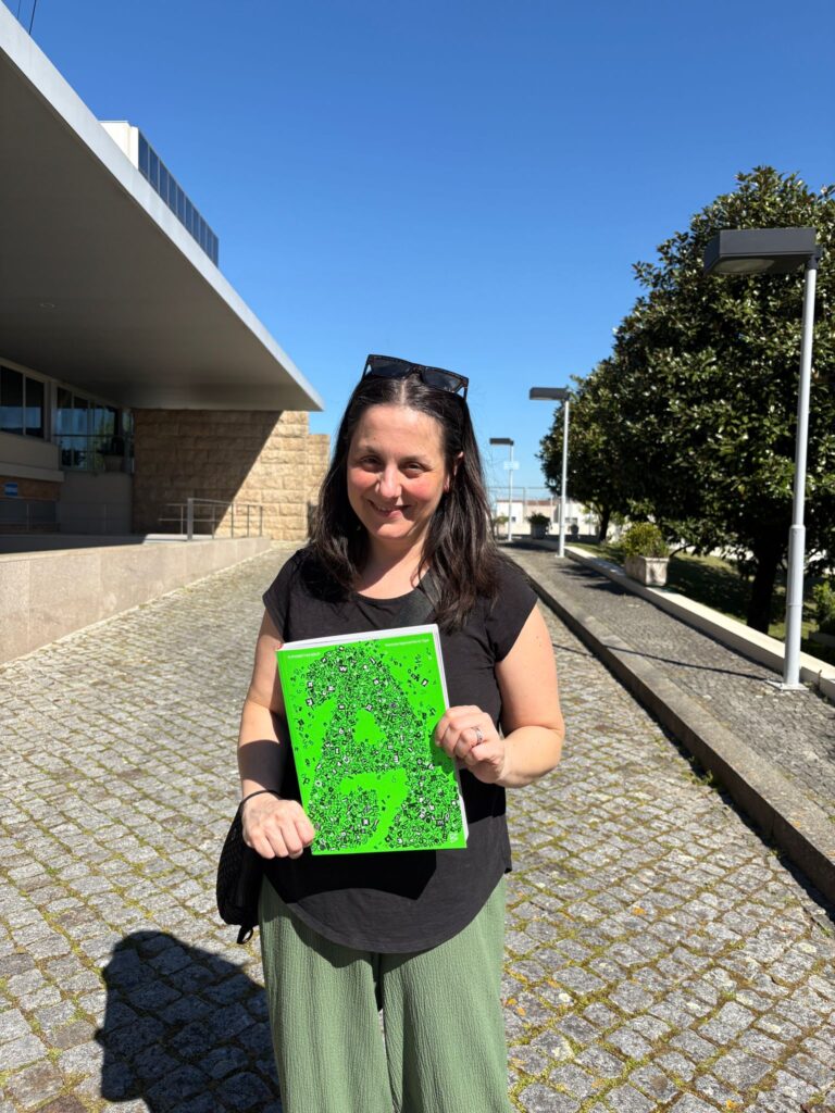a woman smiling and holding a bright large green book in front of a building 