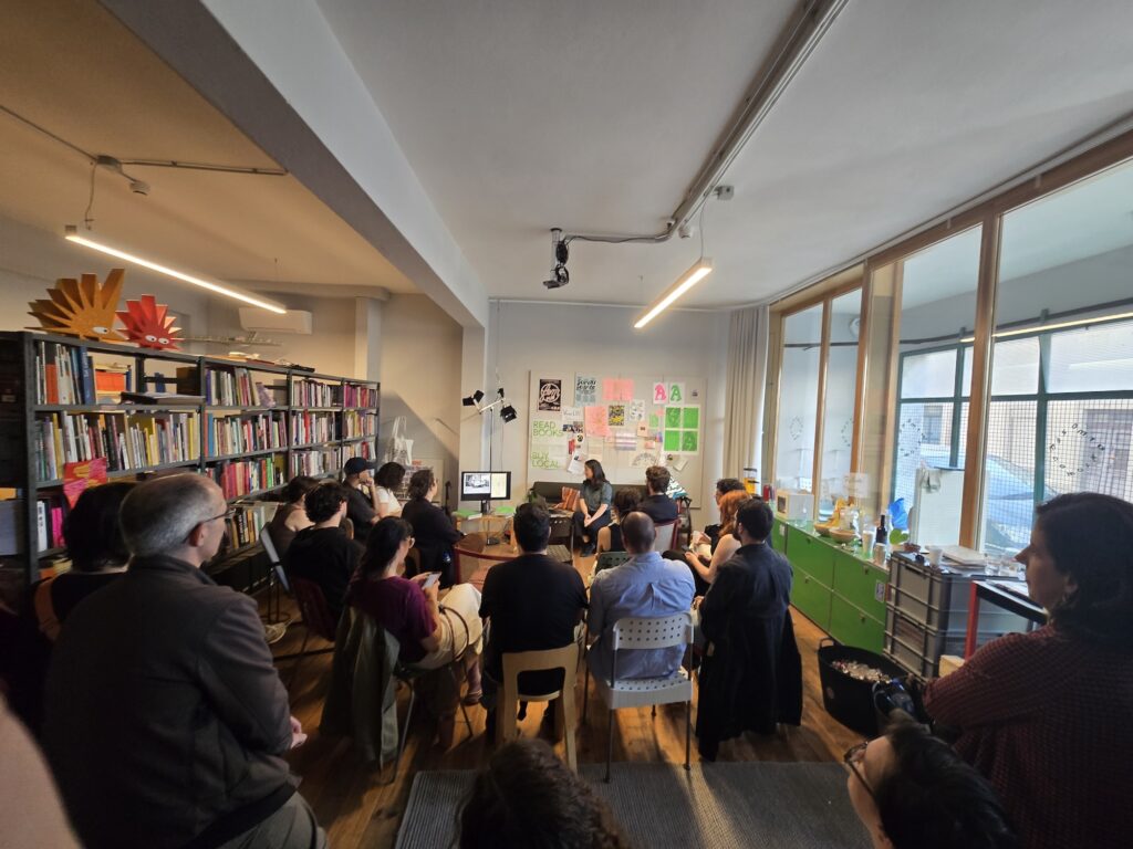a room of people sitting in chairs with a woman at the front of the room sitting next to a monitor giving a talk